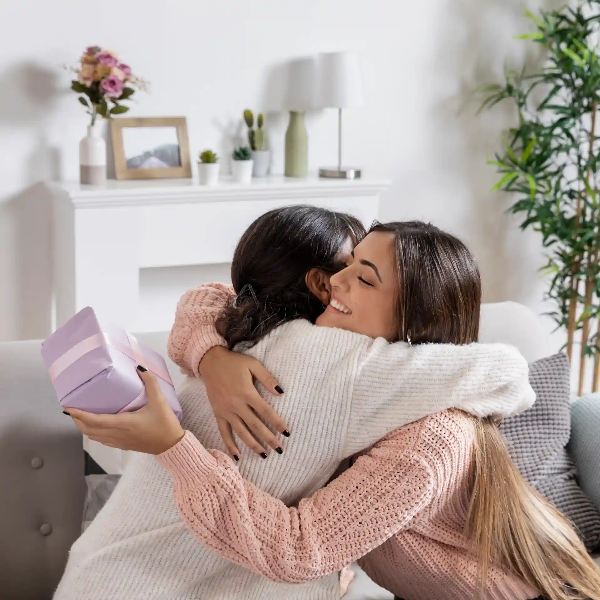 Duas amigas se abraçando, uma delas segurando um presente roxo, em um ambiente aconchegante e iluminado. Transmite amor e carinho.