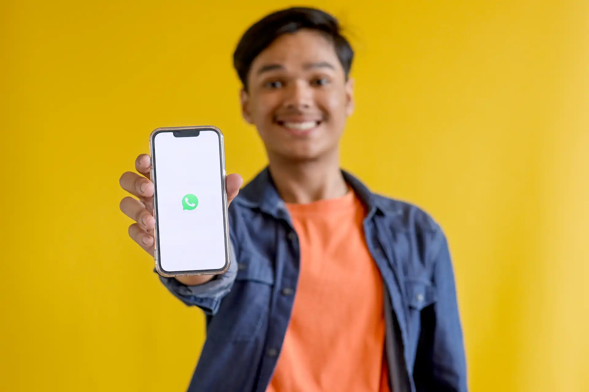 Jovem sorridente segurando um smartphone com o logo do WhatsApp em uma tela branca, com fundo amarelo vibrante. Ideal para conteúdos sobre comunicação e tecnologia.