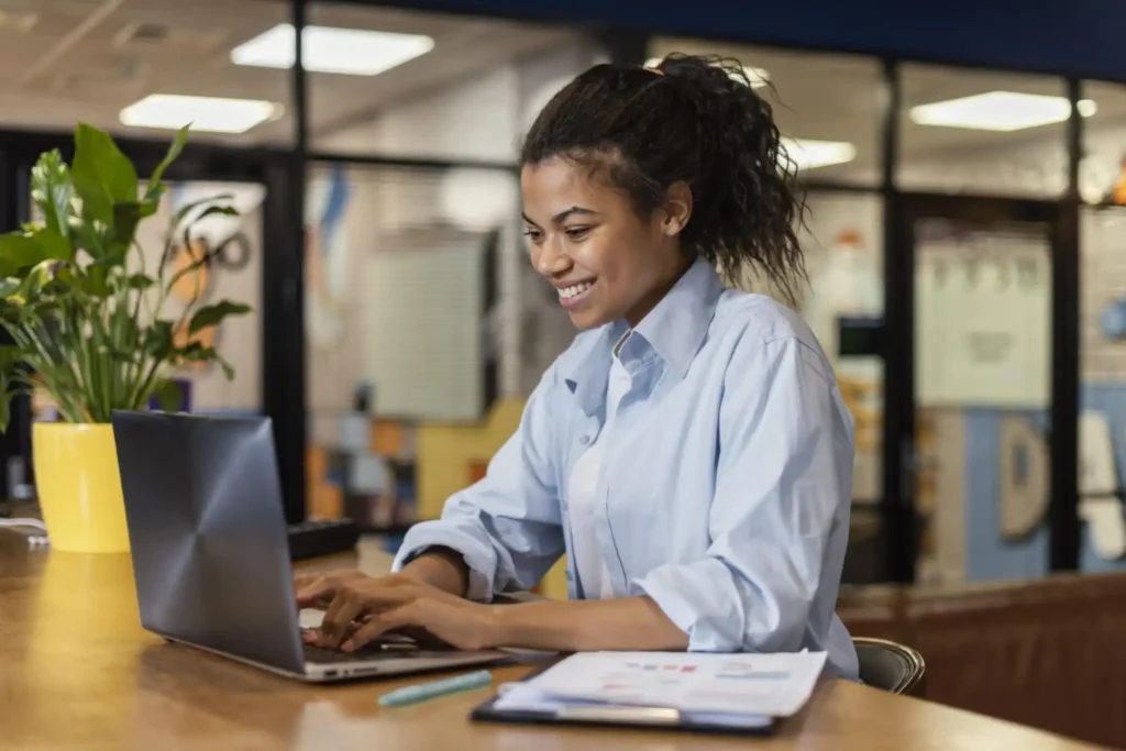 Mulher sorridente trabalhando em um laptop em um ambiente de escritório moderno, com plantas ao fundo, representando produtividade e trabalho remoto.