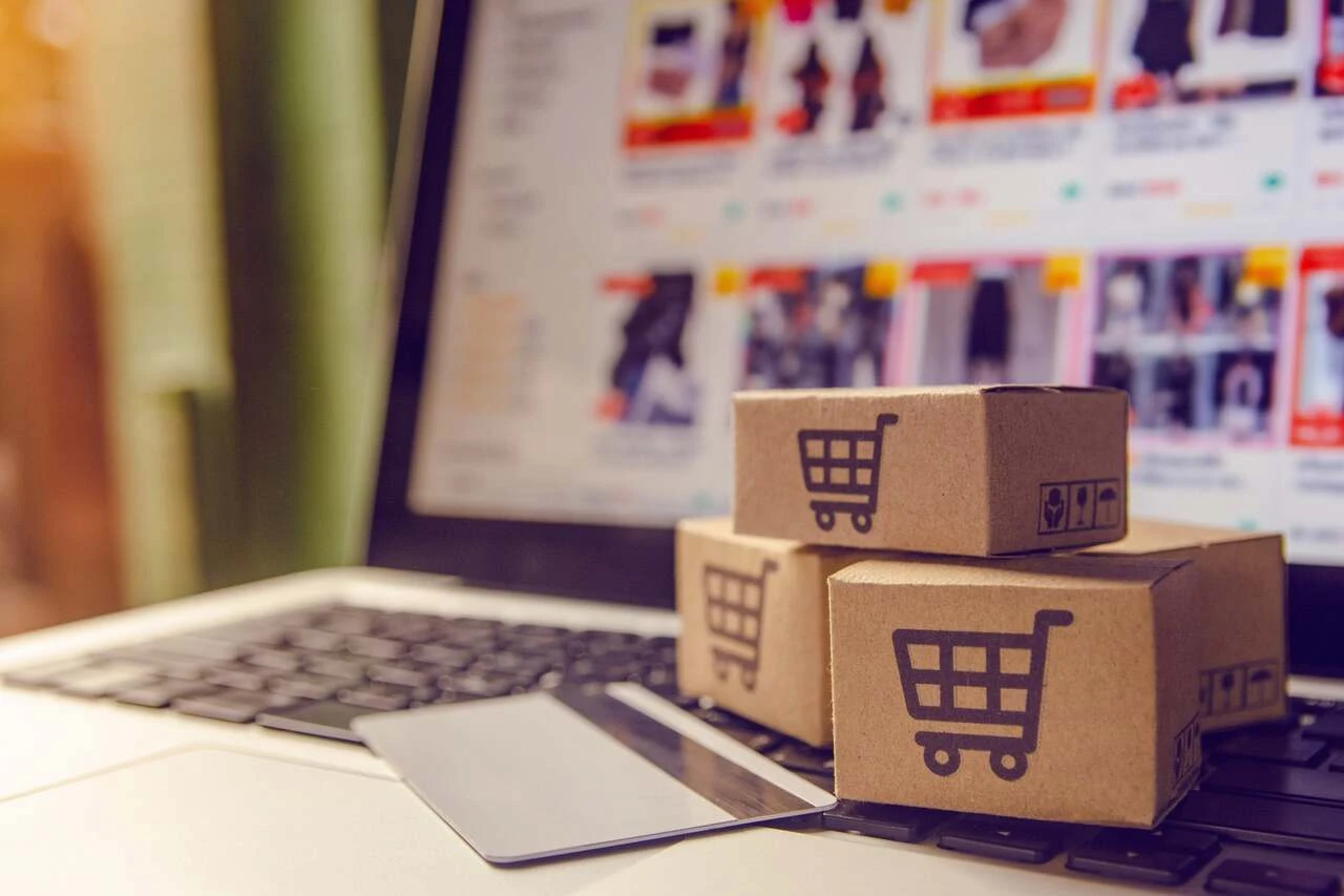 Imagem mostrando três caixas de papelão com ícones de carrinho de compras em cima de um notebook com tela de e-commerce, ideal para ilustrar compras online.