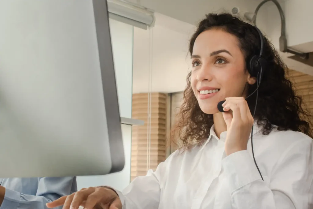 Uma profissional sorridente usando um headset, trabalhando em um computador, em um ambiente de escritório moderno, com boa iluminação e foco em atendimento ao cliente.