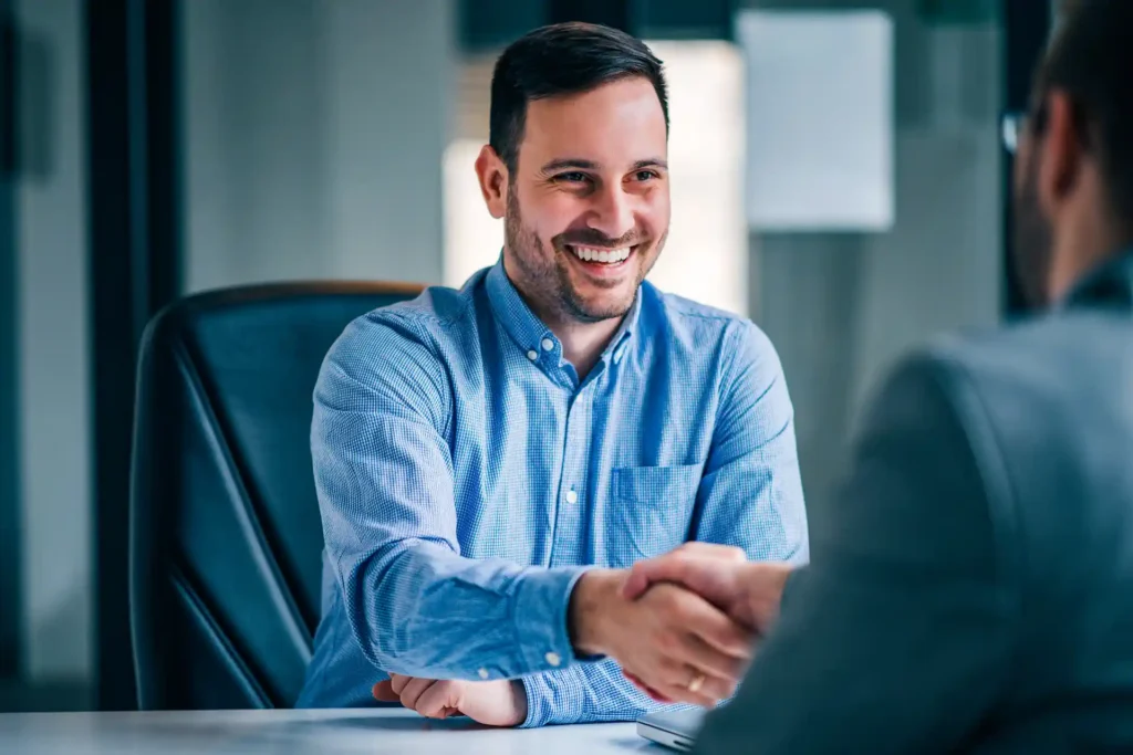 Imagem que mostra um homem sorridente em uma reunião de negócios, fazendo um aperto de mão durante uma negociação amistosa.